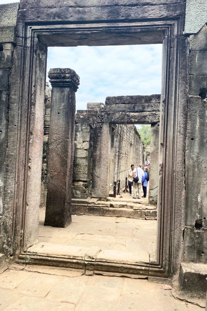 Looking through stone doorframes at a column and a narrow corridor where a guide and two visitors stand.
