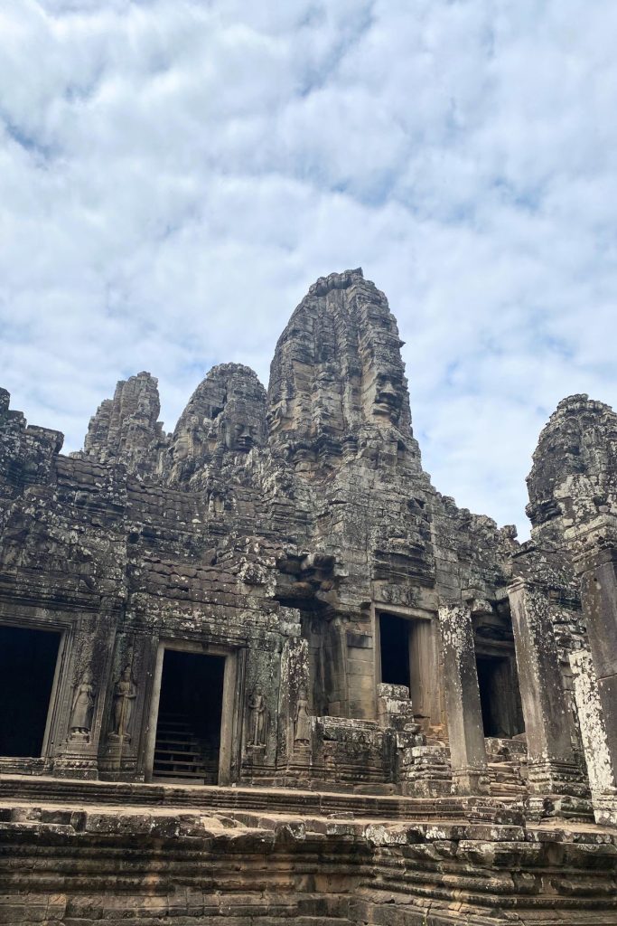 Bayon’s central towers with carved faces above lower rooms with dark doorways, under a cloudy sky.
