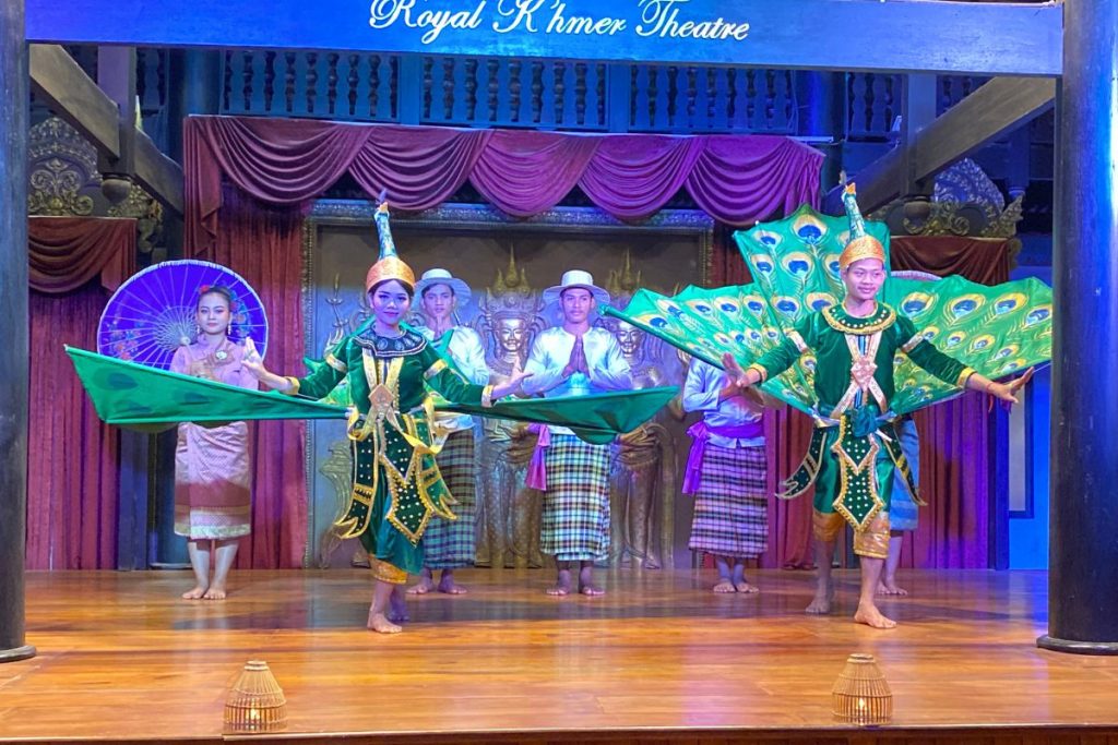 Dancers in green and gold costumes spread peacock-feather wings across a stage beneath a Royal Khmer Theatre sign, with a woman holding a purple parasol behind them.