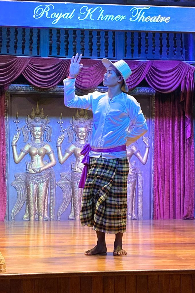 Barefoot man in a white hat, white shirt, and plaid sarong with a purple sash, one hand raised, before golden apsara panels.