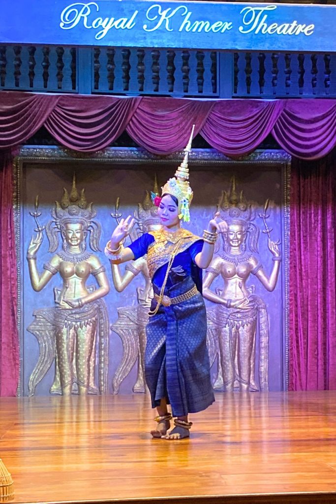 Dancer in a tall gold crown and blue and gold costume with raised hands before three golden apsara figures.