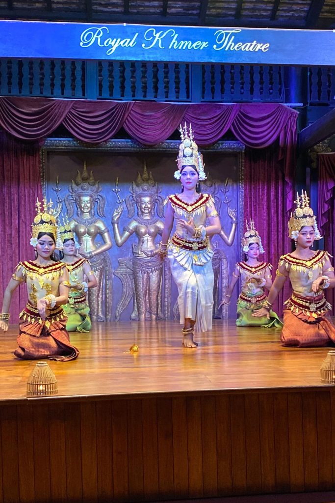 Five dancers in gold-trimmed costumes and tall crowns, one standing at center and the others kneeling, before golden apsara panels.