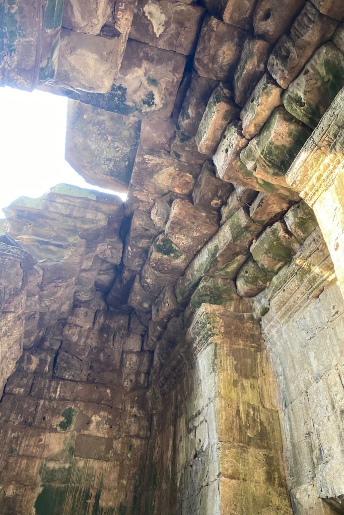 Looking up inside a stone chamber at layered blocks and a gap of bright sky where part of the roof is missing.