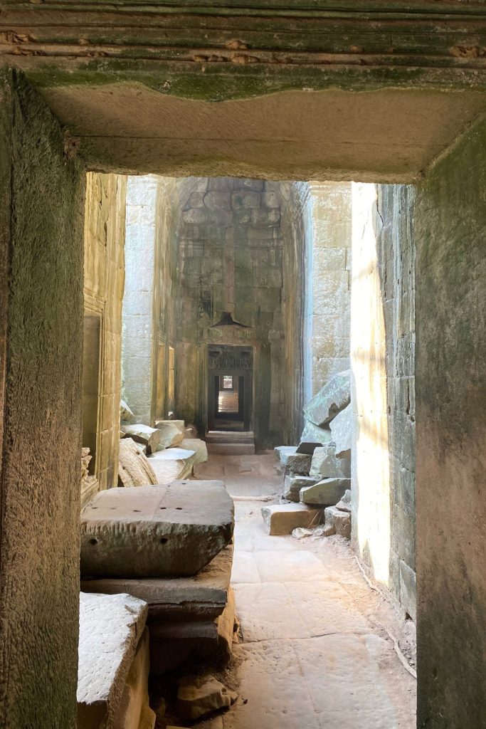Sunlit stone corridor with fallen blocks on both sides and a series of doorways leading away.
