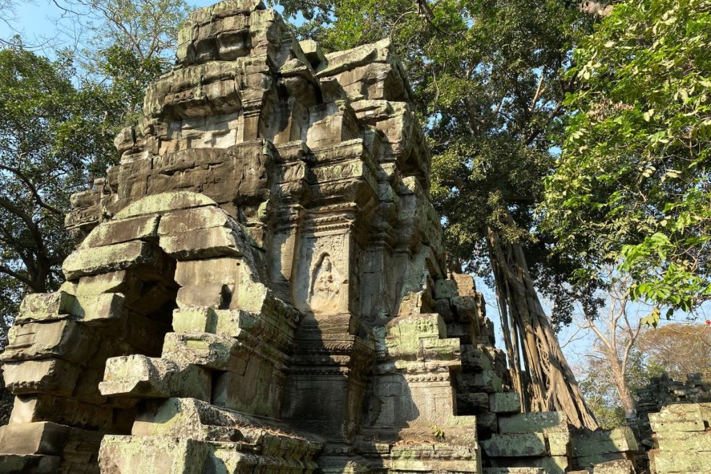 Weathered stone tower at Ta Prohm with a carved niche, trees alongside, and a blue sky behind.