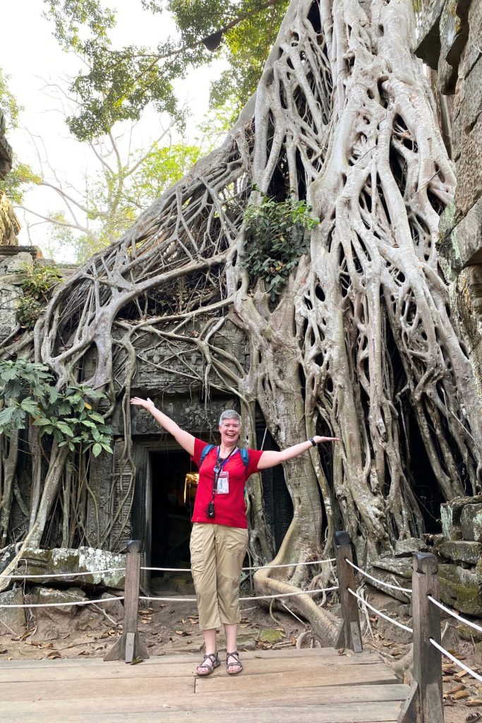 Woman in a red shirt with arms outstretched on a wooden walkway in front of a doorway swallowed by enormous tree roots.
