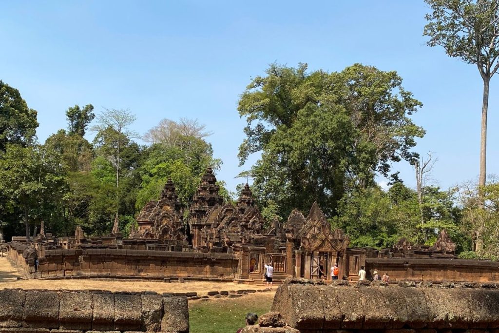 Low red sandstone temple buildings and towers behind a laterite wall, with visitors near an entrance and tall trees behind.
