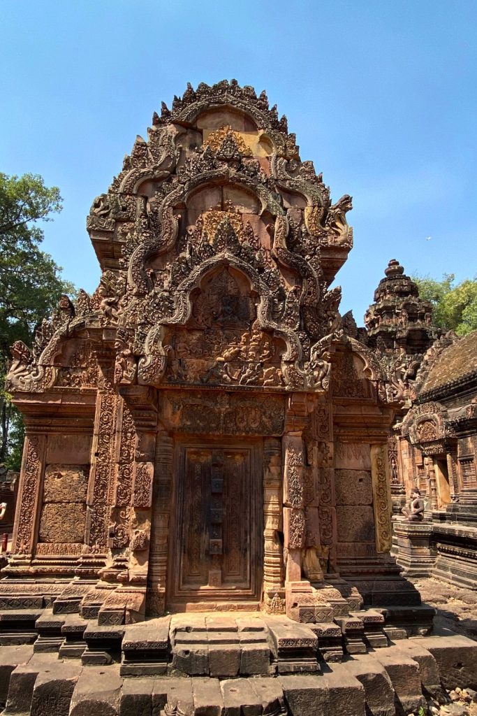 Front of a red sandstone shrine with a carved pediment above a small wooden door and carved pilasters on both sides.