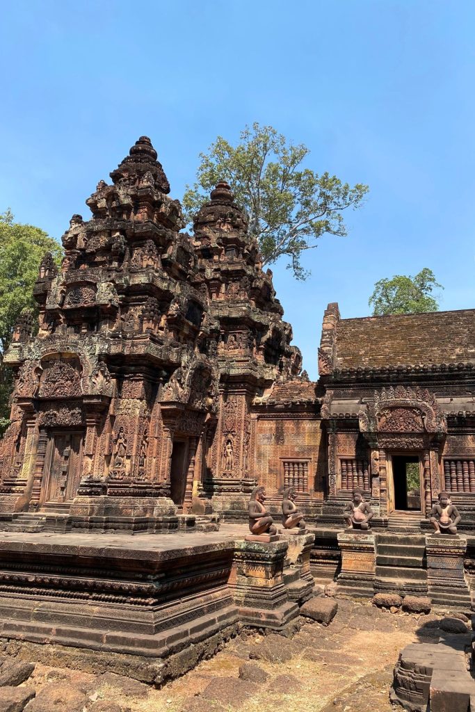 Two red sandstone towers with carved niches and kneeling stone guardian figures in front, beside a tiled-roof building.