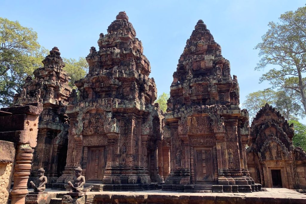 Two stepped red sandstone towers covered in carvings, with kneeling guardian figures on the platform in front and tall trees behind.