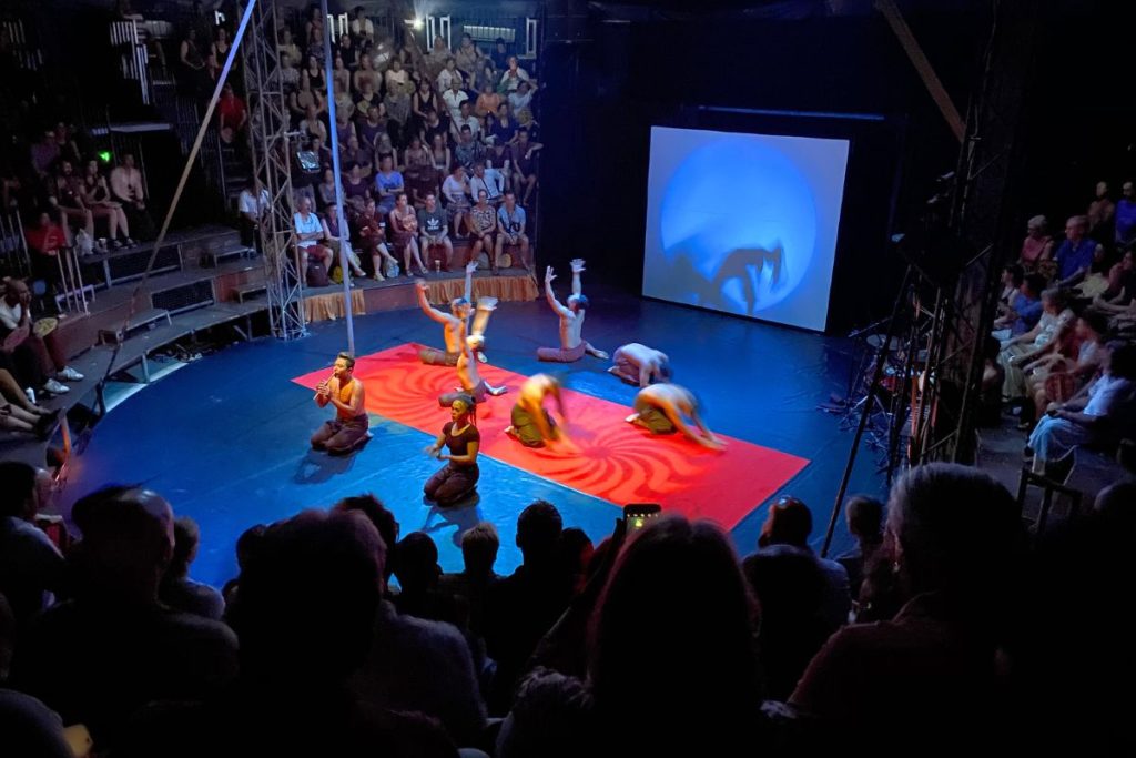 Performers kneeling and stretching on a red mat in a circular arena, with tiered audience seating around them and a blue projection screen behind.