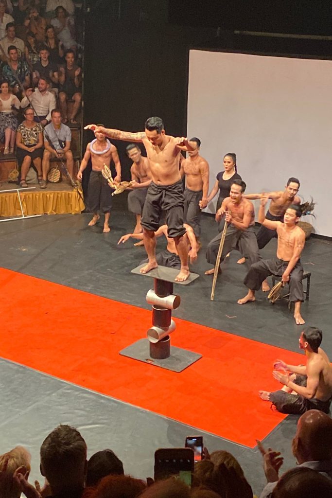 Barefoot performer with arms out balancing on a board atop stacked metal cylinders, while others watch from the red mat.