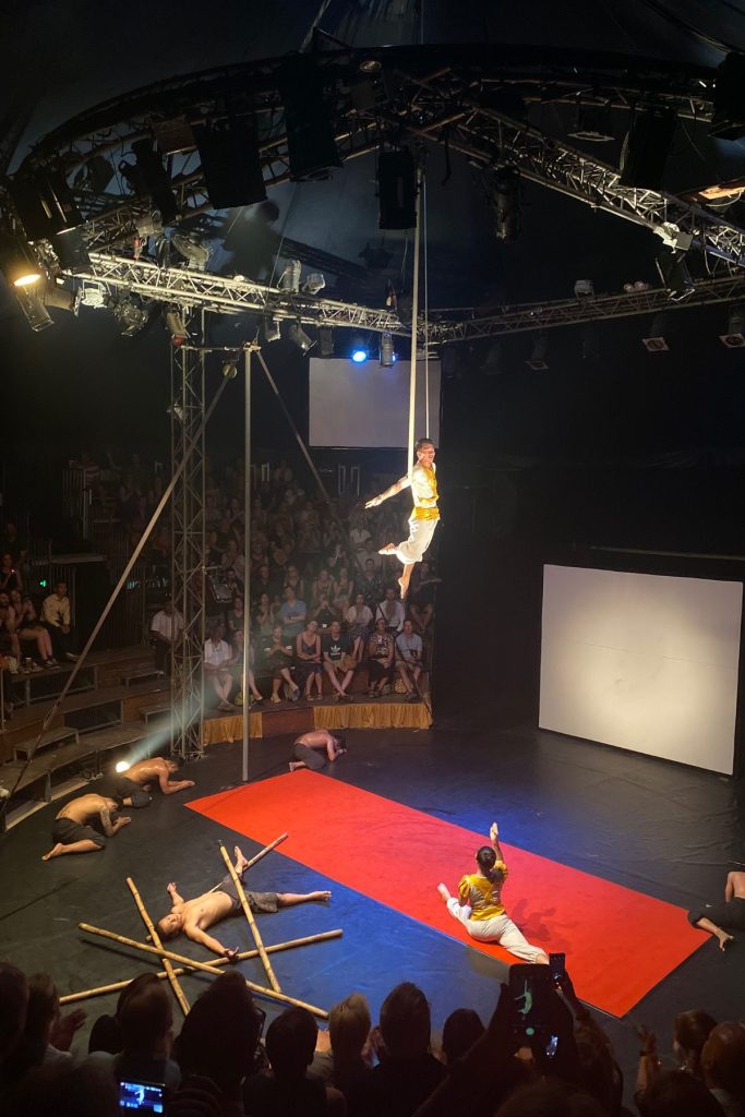 Performer in white and yellow hanging from a rope high above the stage, with others on the floor below and the audience watching.