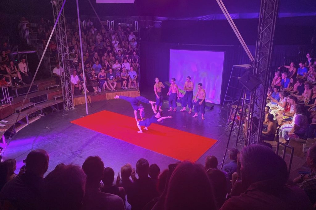 Two performers in blue balancing in an acrobatic pose on a red mat while four performers stand behind them and the audience looks on.