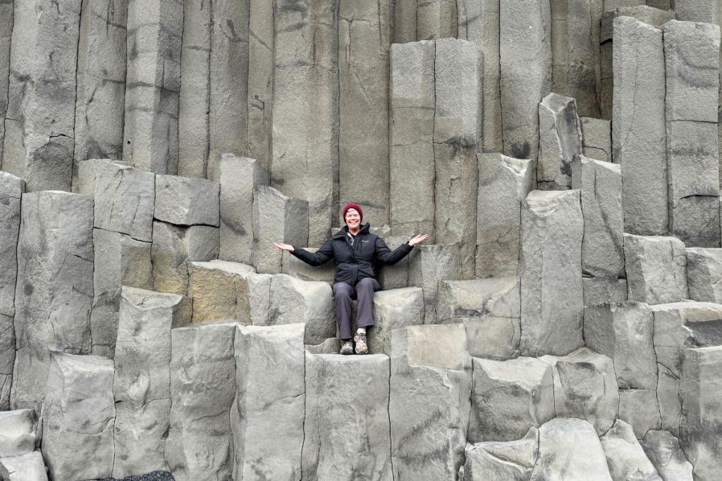 A smiling woman, wearing a black jacket, gray pants, and a maroon beanie hat, sits amongst a large gray rock formation at Reynisfjara black sand beach, Iceland