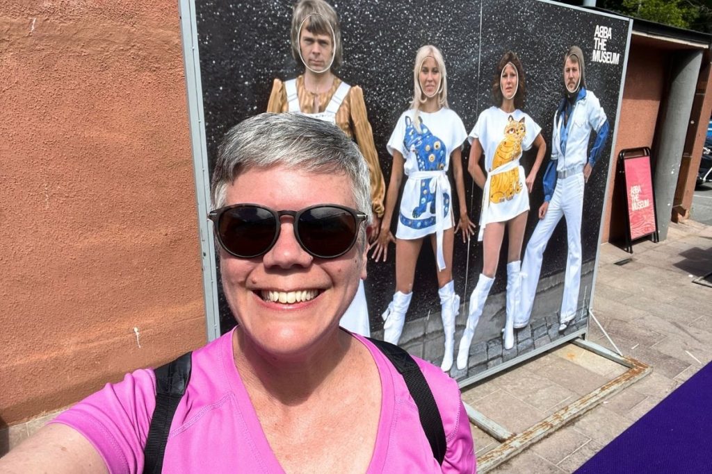 A smiling woman, wearing a pink shirt and dark sunglasses, stands in front of a large photo of the four members of the band ABBA, in front of the ABBA Museum in Stockholm