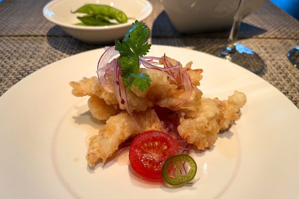 A white plate is artfully arranged with a pile of crispy golden pieces of lobster tempura. One slice of red tomato and one slice of jalapeño pepper sit in front of the lobster, and thin pieces of sliced red onion and a sprig of cilantro sit atop the lobster. A white bowl with several pieces of edamame is behind the plate of lobster.