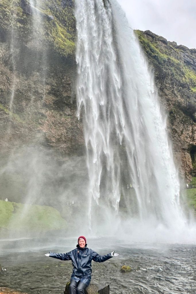 A smiling woman, wearing black rain jacket and pants and a maroon beanie hat, sits on a rock in front of an active waterfall, her arms are outstretched perpendicular to her body, with her palms facing up. 