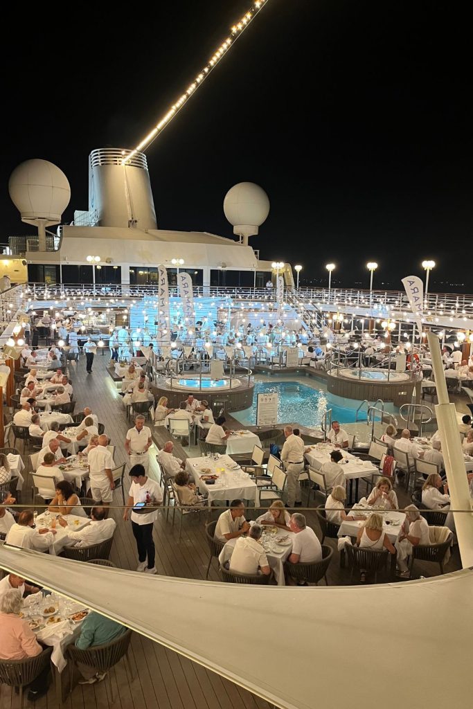 A large group of passengers, all dressed in white, sit at many different tables arranged around a pool on a cruise ship at night.