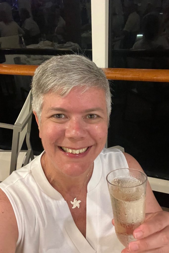 A smiling woman, wearing a white sleeveless top and a necklace with a small white starfish, raises a glass of champagne in a toast towards the camera. She is sitting on a cruise ship pool deck, with the black night sky visible in the windows behind her.