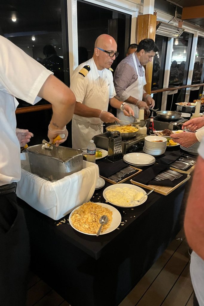 A bald man, wearing eye glasses and a white ship officer's uniform and apron, is spooning food from the skillet in front of him onto a plate someone is holding out to him. Other men are next to him, also serving food.