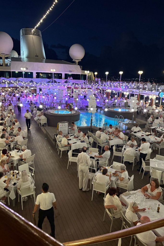 A large group of passengers, all dressed in white, sit at many different tables arranged around a pool on a cruise ship at night.