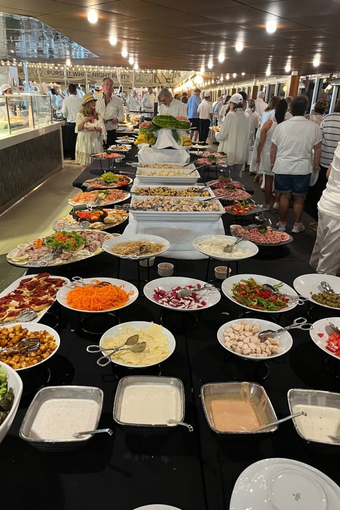 A large buffet table on a cruise ship's pool deck. Several white plates of food are sitting on black tablecloth-covered tables. A crowd of people wearing white are walking around the table.