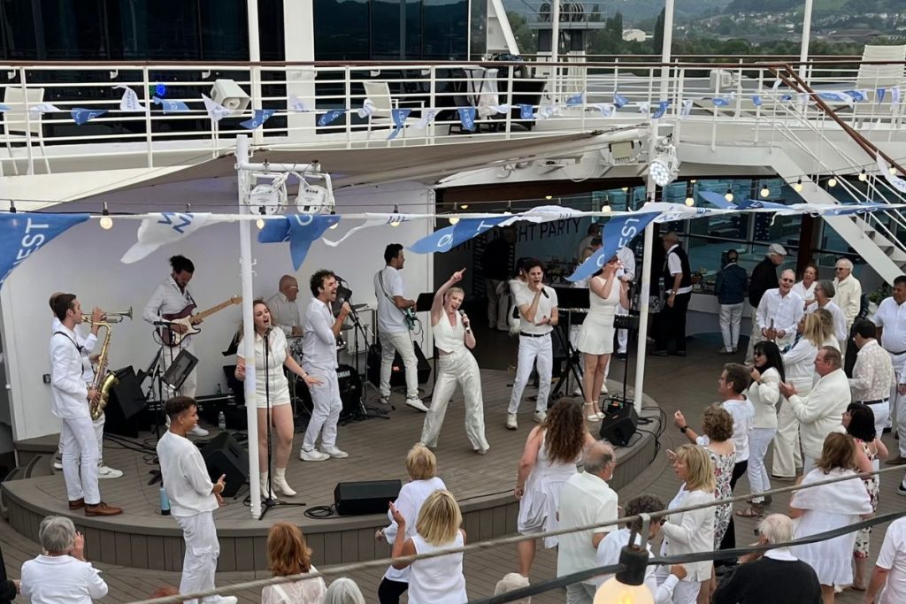 A group of nine performers, all dressed in white, are singing and dancing on the stage on a cruise ship's pool deck. A crowd of people, also dressed in white, are dancing on the dance floor in front of the stage.
