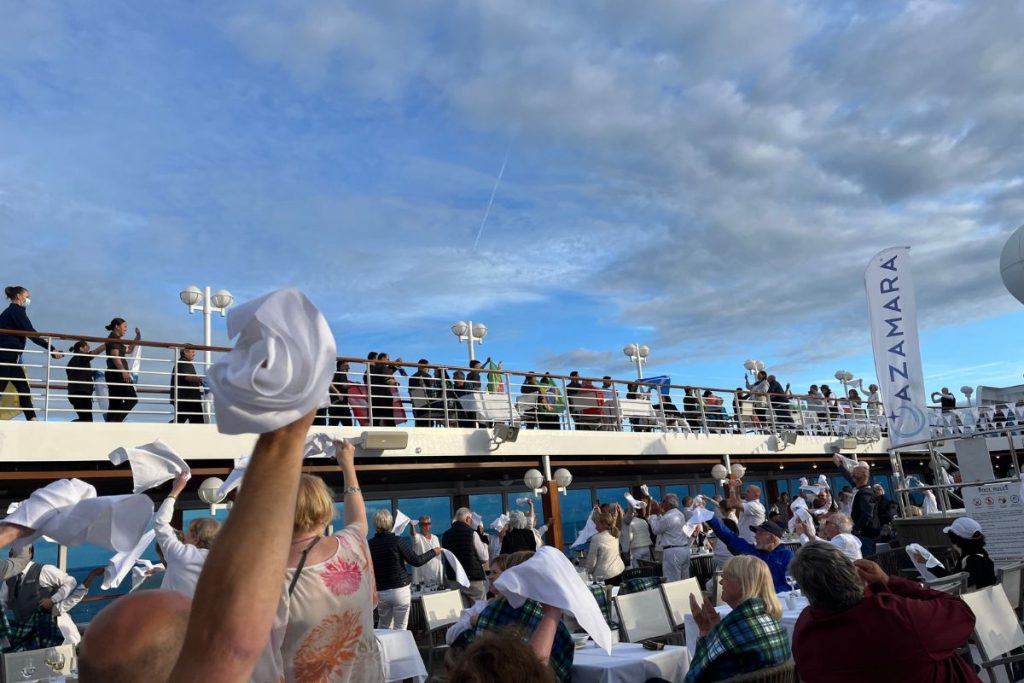 A large crowd of people are sitting at tables on a cruise ship's pool deck, with more people walking on a jogging track above the pool deck. Many people in the crowd are waving white napkins above their heads.