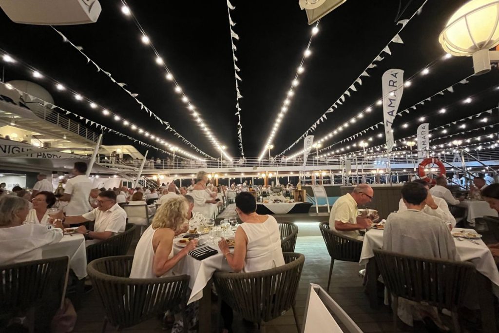 Passengers dressed in white are sitting and eating at several tables on a cruise ship's pool deck, with strings of white lights spanning the upper deck area.