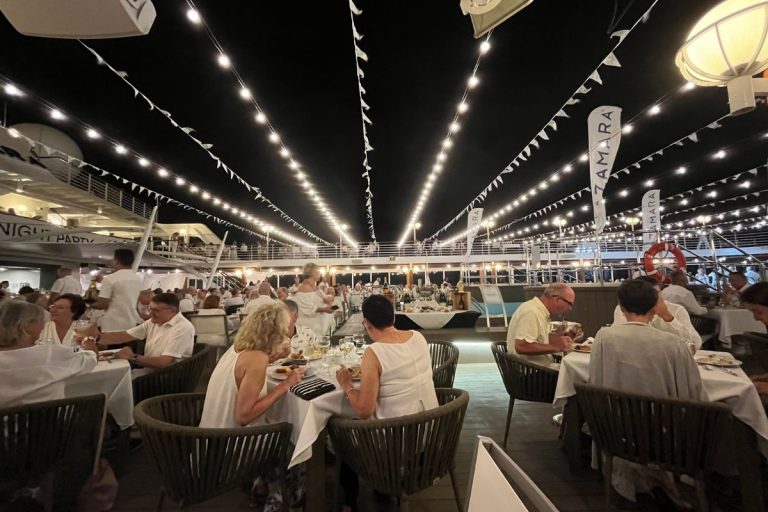 Passengers dressed in white are sitting at several tables on a cruise ship's pool deck, with strings of white lights spanning the upper deck area.