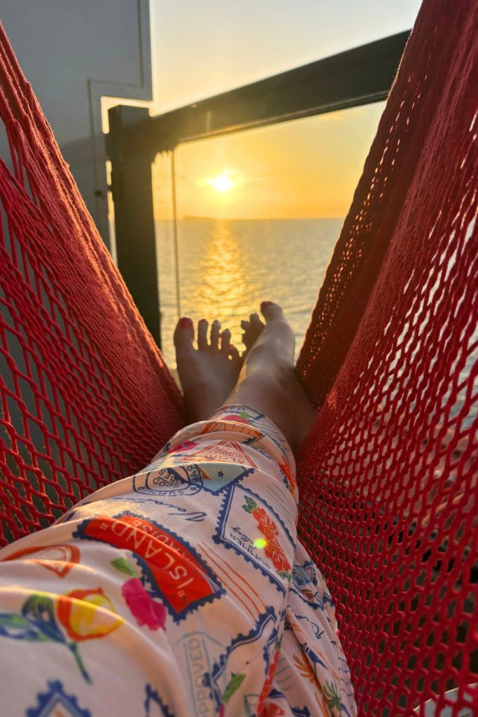 A woman's legs, dressed in brightly colored pants, are crossed and are lying on a red mesh hammock on the balcony of a cruise ship cabin, with the sun setting over the water in the background