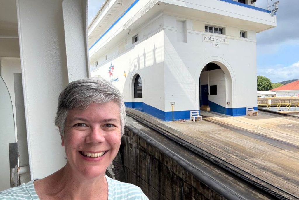 A smiling woman, wearing a mint green and white striped shirt, stands on a cruise ship balcony, with the Pedro Miguel lock on the Panama Canal in the background