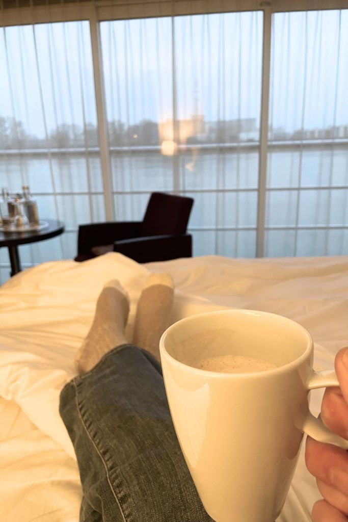 Legs, dressed in blue jeans and brown socks, are crossed and laying on top of a bed covered with a white comforter. In the foreground, a hand holds a white mug filled with a foamy liquid. In the background, a large series of sliding glass doors is covered with a filmy curtain, with a view of a river and some buildings on the opposite river bank. A black arm chair and a side table with some bottles is set in front of the sliding glass doors.