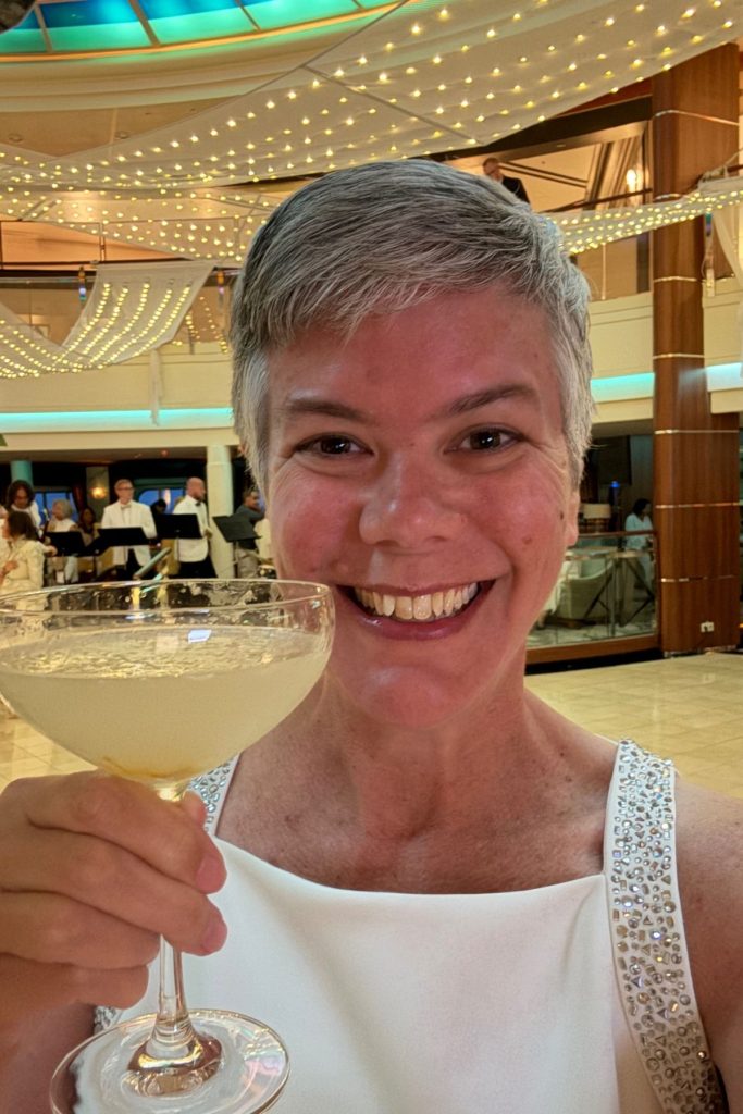 A smiling woman, wearing a white sleeveless dress with rhinestone embellishments on the top, raises a glass of a pale colored drink in a toast towards the camera. She is standing in an atrium on a cruise ship, with festive white decor and lights strung from the ceiling behind her.