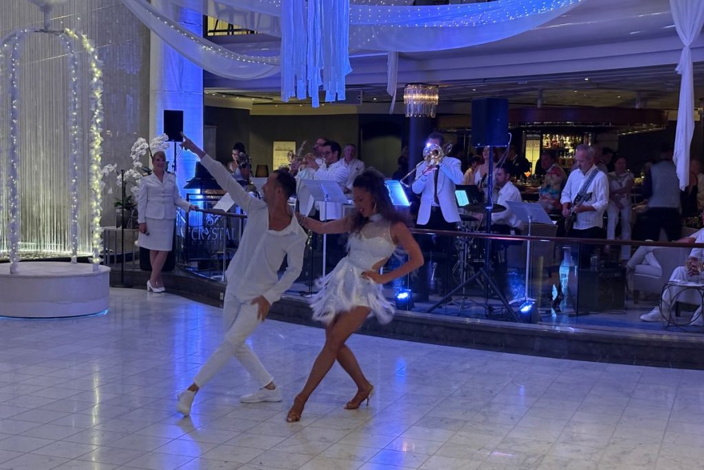A man and a woman, both dressed in white, perform a dance on a white marble floor in a cruse ship atrium. A band, wearing white dinner jackets and black pants, performs behind them.