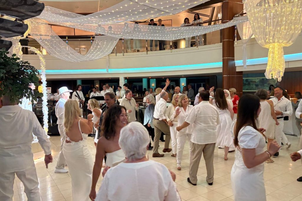 A large group of people, all dressed in white, dance on a white marble dance floor in the atrium of a cruise ship. Festive white decorations and lights are strung above the atrium.