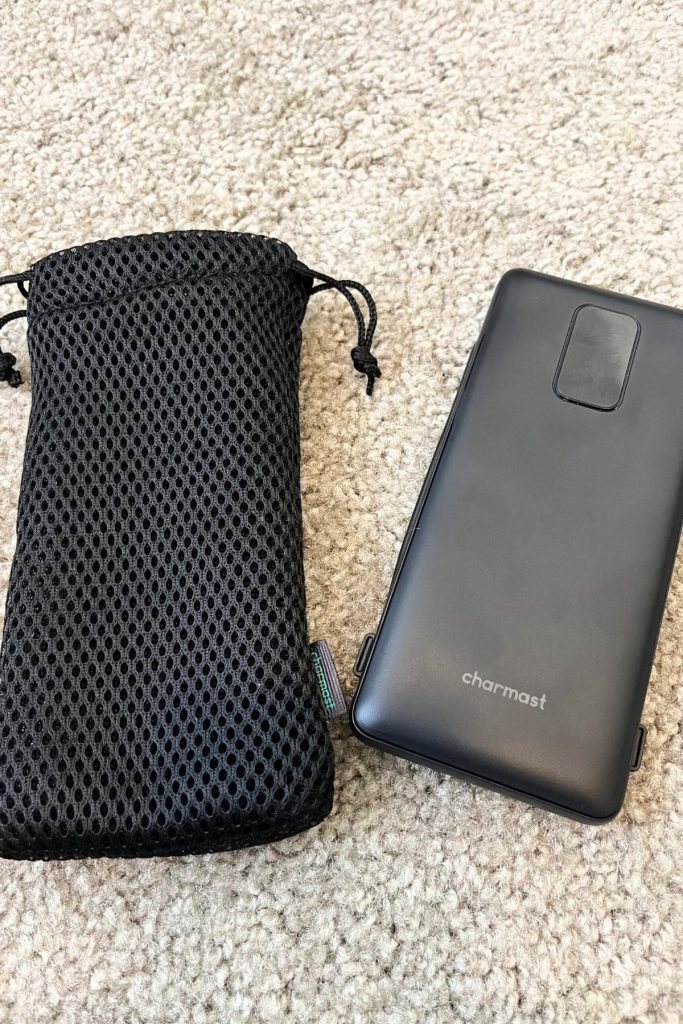 A black portable power bank is on the right, with its black mesh carrying case on the left. The power bank has the word Charmast in gray near the bottom.