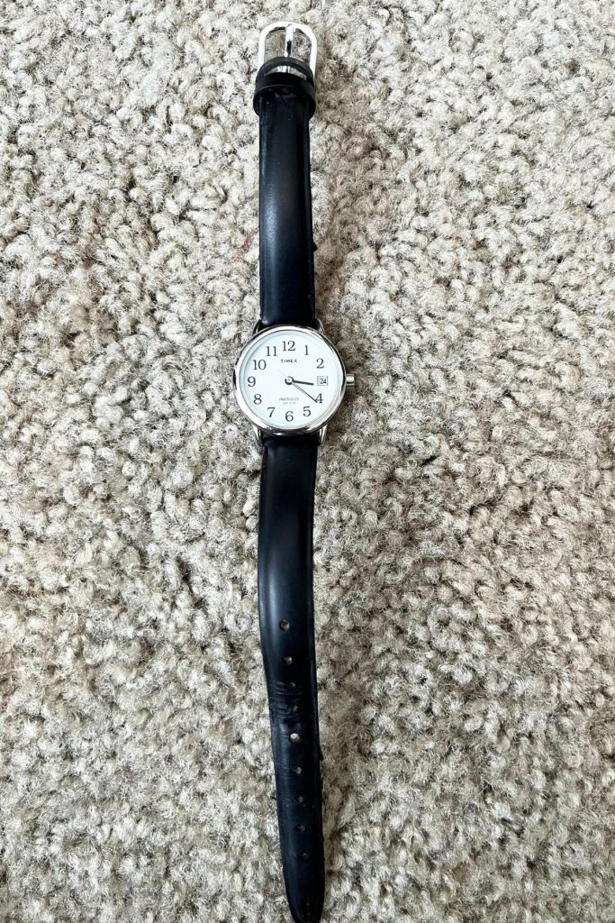 A woman's wristwatch, with a black leather strap, white face, black numbers, and black hands, showing the time of 3:21 and the date of 24.