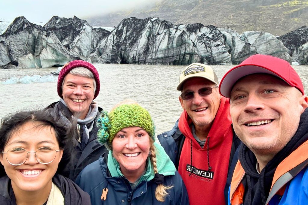 A group of three women and two men, all smiling, stand in front of a body of water with a series of glaciers behind the water.