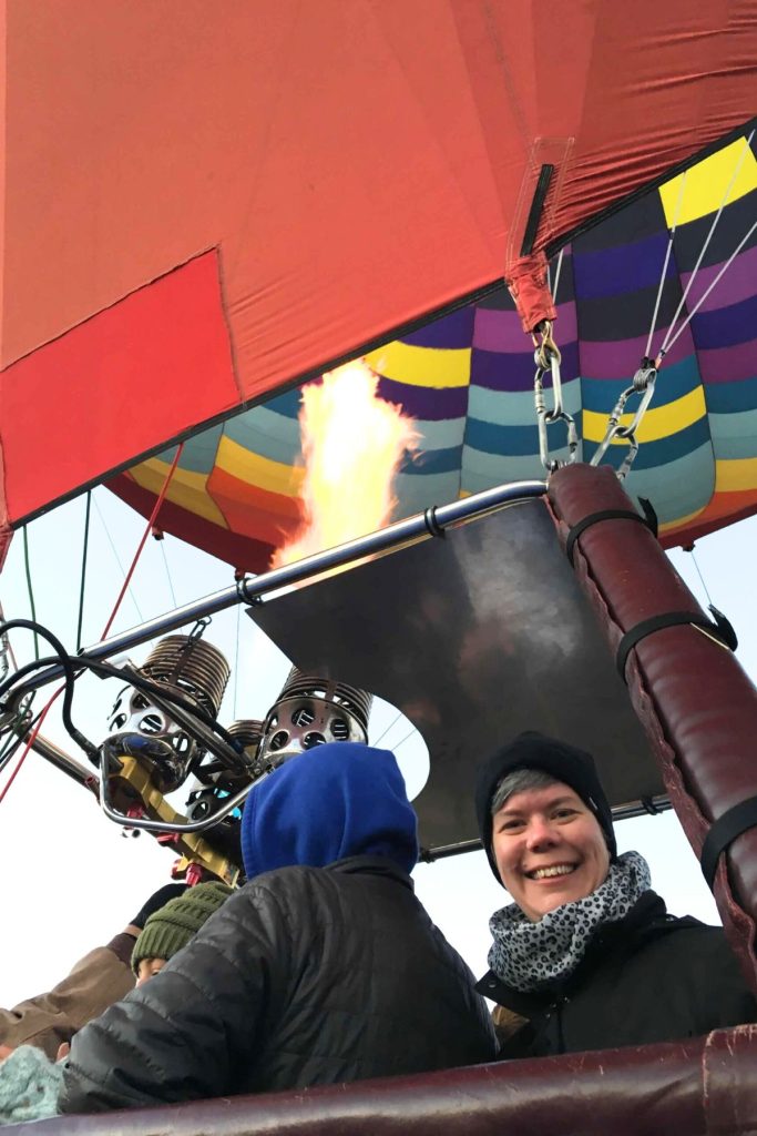 A smiling woman, wearing a black jacket, a black beanie hat, and a black and gray leopard print scarf, stands in the basket of a hot air balloon with other people. The balloon is brightly colored, and a large flame is blowing up into the balloon.