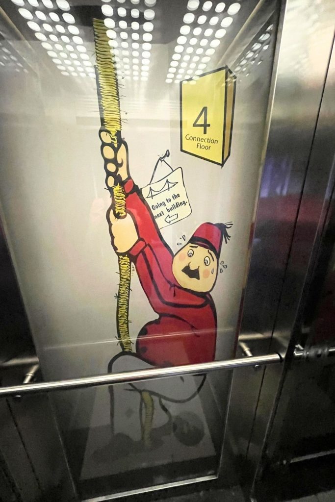 A humorous cartoon drawing of a man dressed as a hotel bellhop gripping a rope hanging from the ceiling, with signs behind him that read Access to the Next Building and a large number 4 with the words Connection Floor below it.