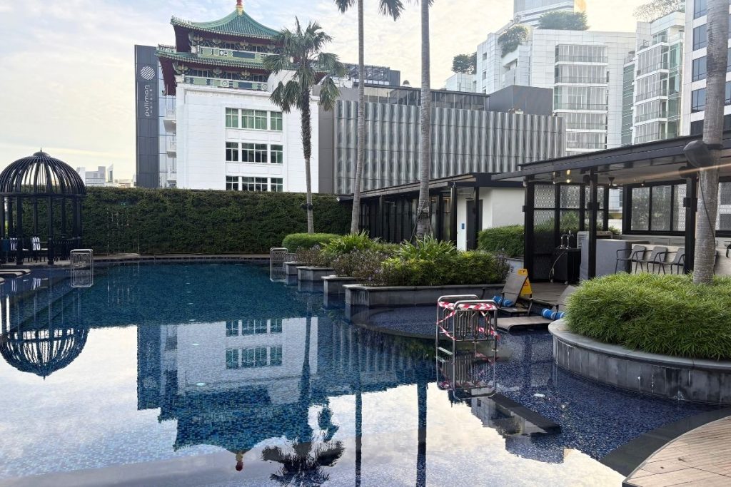 A tranquil and still hotel pool, with cabana areas on the right side and large city buildings in the background