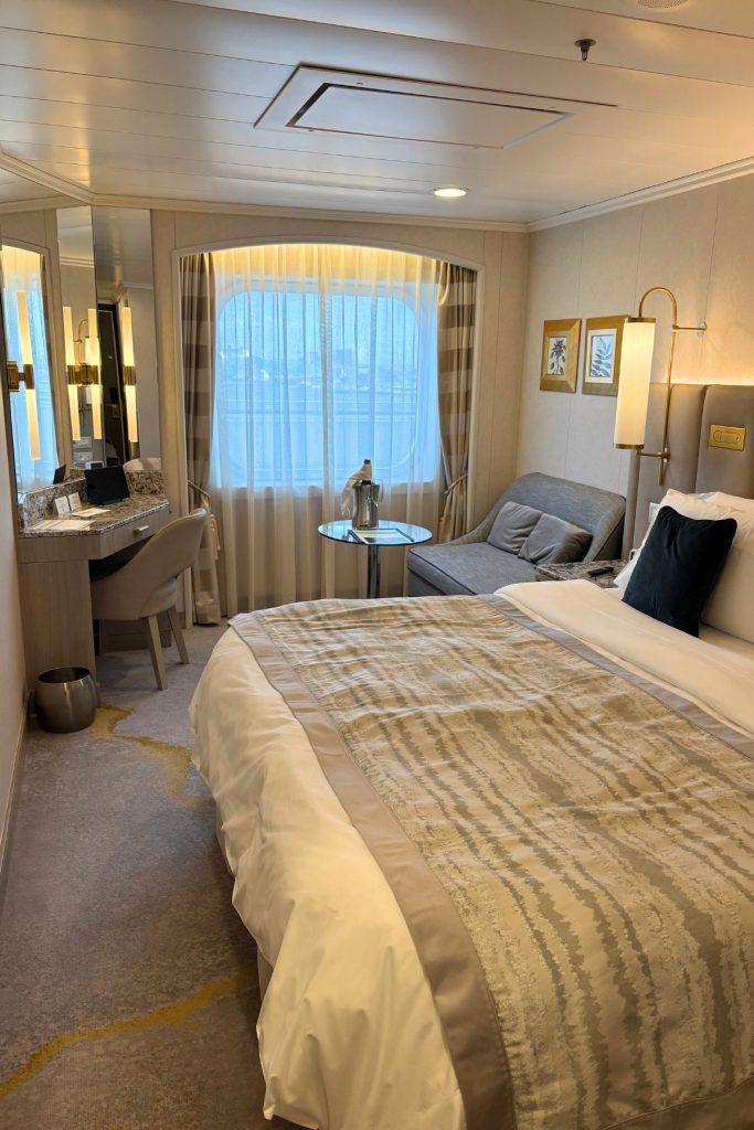 Looking into an ocean view cruise ship cabin on a Crystal ship. A large picture window is centered on the far wall, covered with sheer drapes. A seating area with a gray couch, glass-topped table, desk, and arm chair are in front of the window. A large bed is seen in the foreground.