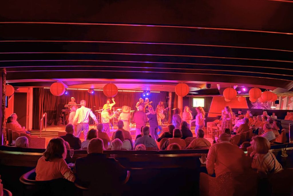 A group of people sit in the audience, all watching a band perform on the stage. Several people are dancing on a dance floor between the audience and the band.