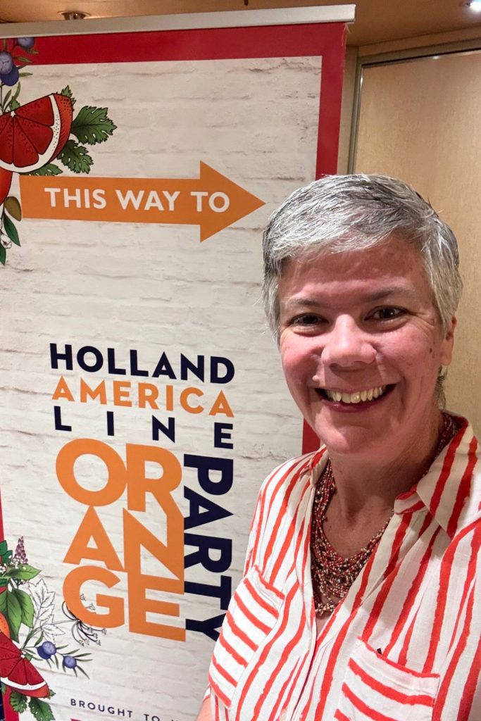 A smiling woman, wearing an orange and white striped collared shirt and a beaded necklace, stands in front of a large sign that reads, This way to Holland America Line Orange Party.