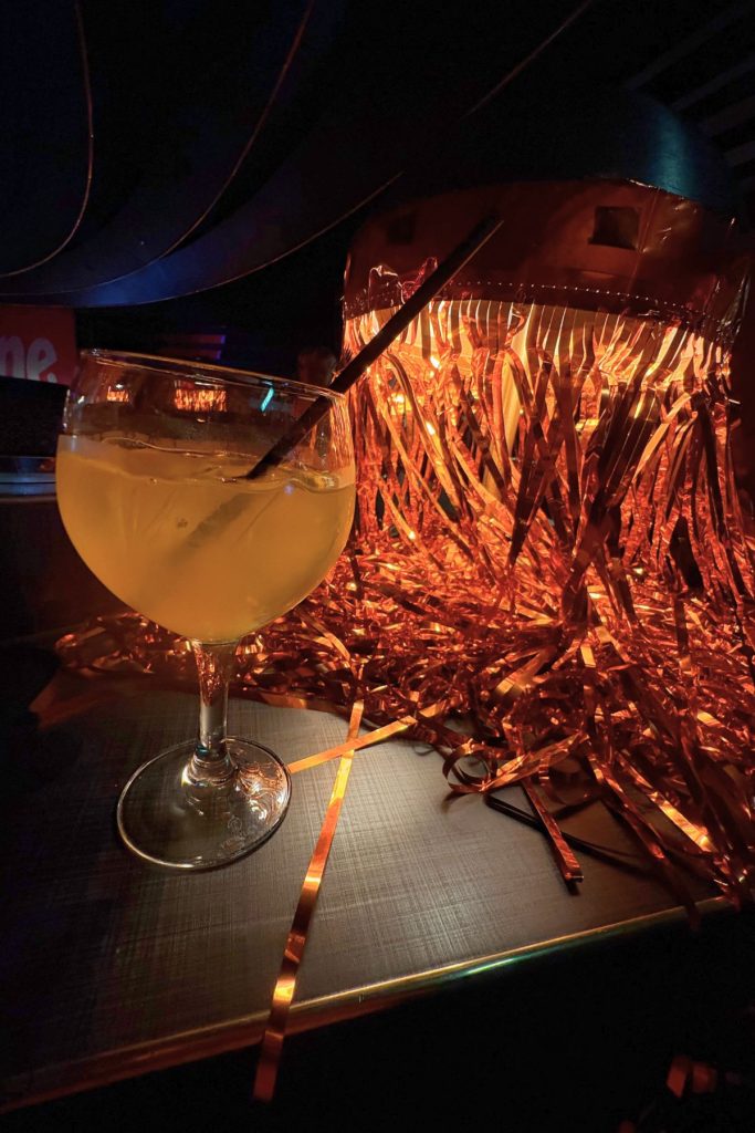 A stemmed glass goblet filled with an orange colored drink is on the left. To the right is a table lamp covered with orange colored streamers. Both are sitting on a dark brown table.