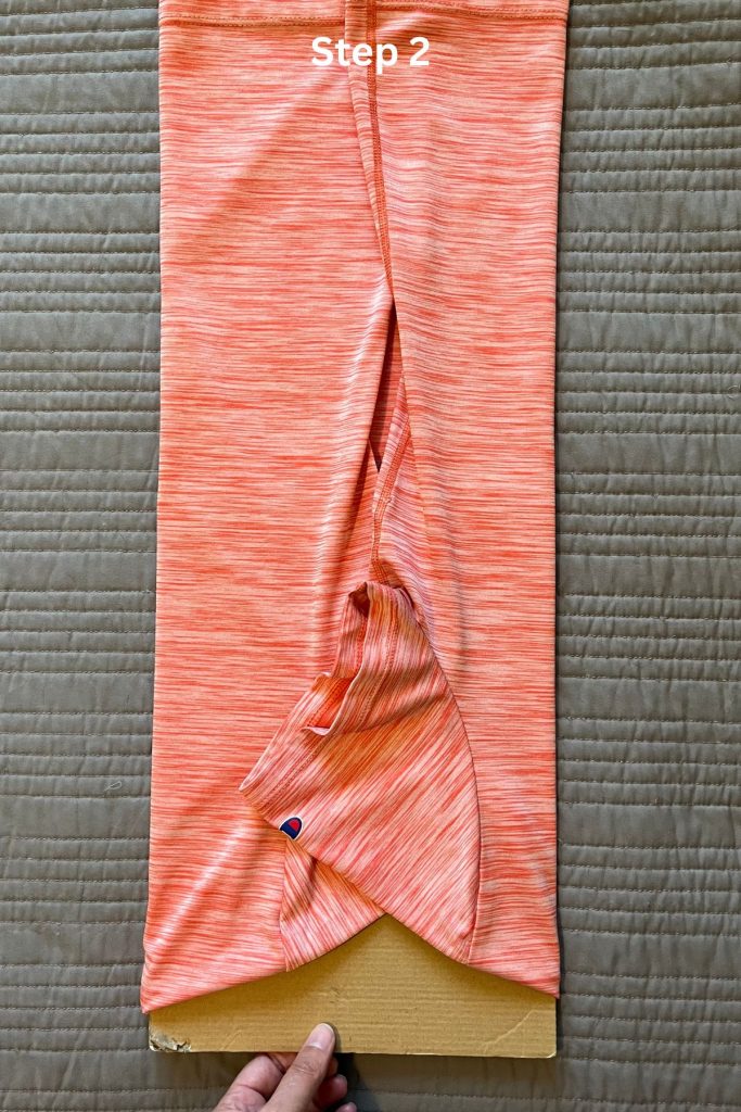 An orange tee shirt is laid out, folded in thirds lengthwise around a smaller rectangular piece of brown cardboard at the neck opening. A hand is pulling the cardboard out from the shirt at the bottom.