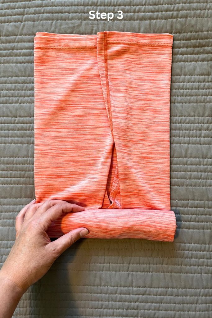 An orange tee shirt is laid out, folded in thirds lengthwise. A hand is rolling up the shirt from the bottom.