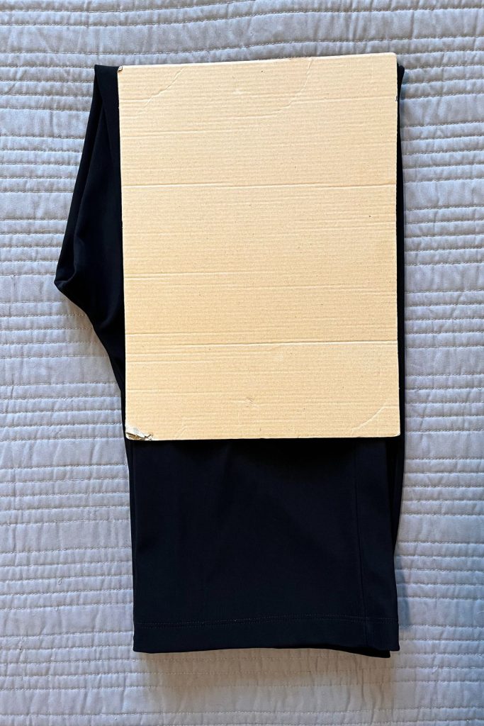 A pair of black pants is laid out, folded in half with a smaller rectangular piece of brown cardboard on top at the fold. Some of the pant legs are still visible at the bottom.